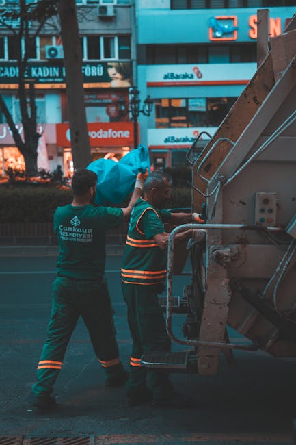 A street scene featuring a recycling or waste collection truck with its rear compartment open, revealing hydraulic mechanisms and a container for waste loads. A worker in high-visibility red and yello