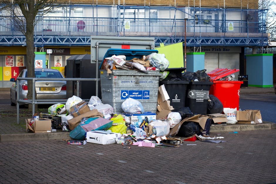 A man wearing a dark jacket, black pants, grey knit cap, and white gloves stands on a modern train station platform, beside a large, red metal trolley heavily loaded with various rubbish bags made of 